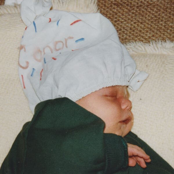 Newborn Conor lying asleep wearing a dark green jumper. On his head is what looks like a cloth nappy which has been repurposed into a hat by his older siblings. On it, they have written “Conor”.