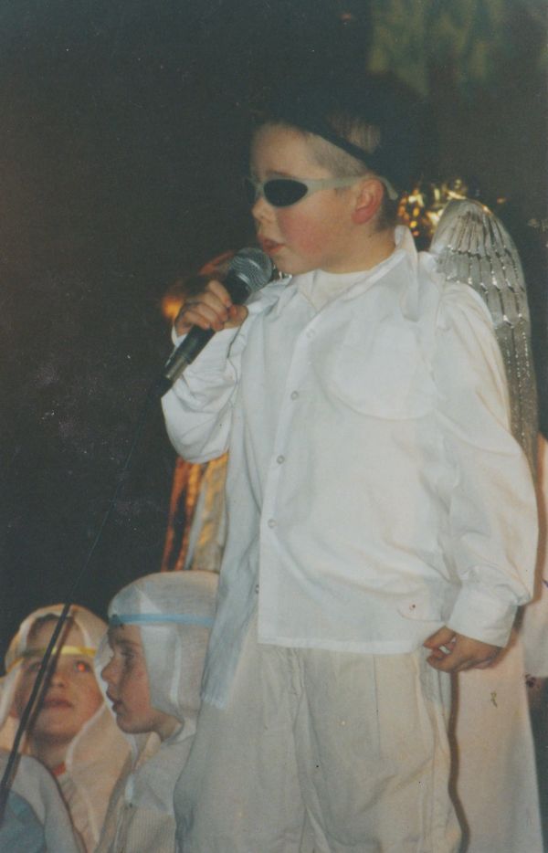 Conor, around age five, playing an angel in his school drama production. He is wearing all white, with silver angel wings on his back, cool-guy sunglasses and has a microphone in his hand held up to his mouth.