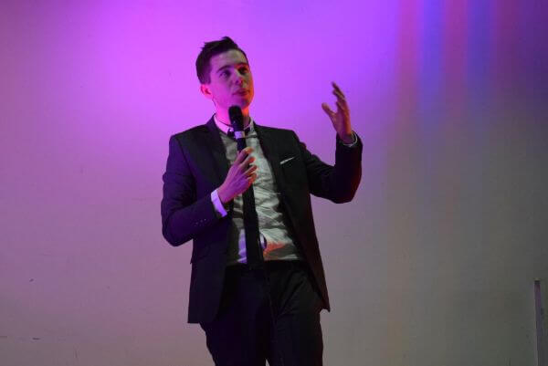 Conor in his early twenties, stood on stage in a suit. He is holding a microphone and performing stand-up. The photo is tinted purple from the stage lighting.