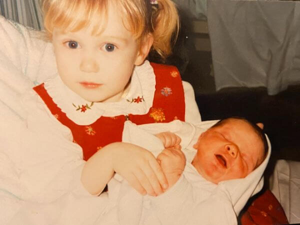 Laura, aged three, holding her newborn brother Daniel for the first time. She is wearing a red pinafore dress over a white blouse, has blonde pigtails and is looking at the camera while holding Daniel delicately. Daniel is wrapped in a white blanket with his eyes closed and his mouth open, asleep.