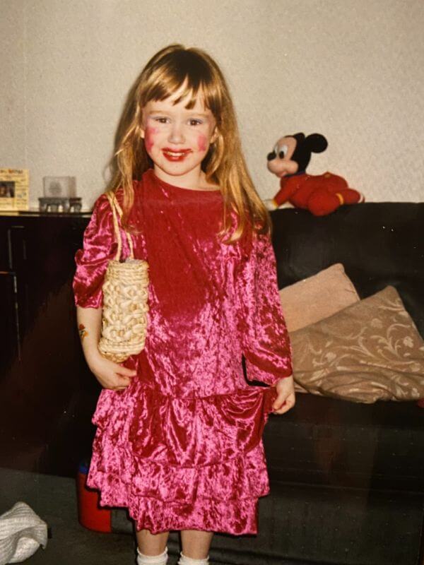 Laura, around age five, stands in her living room. She is wearing a bright pink velvet dress, which she is holding up on one side. On the other arm she has a basket-style handbag over her shoulder. She has clearly done her own make-up — there is blue eye-shadow smeared on her eyelids, messy red lipstick around her mouth and two bright pink circles painted onto her cheeks.