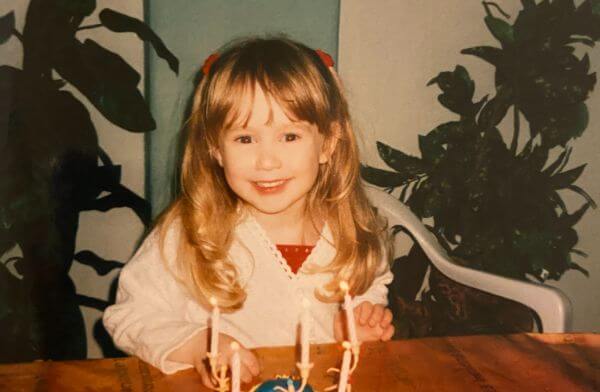 Laura at her fifth birthday party. She is sitting in a white blouse in front of a birthday cake with five candles. Her blonde hair falls around her shoulders and she is smiling widely.