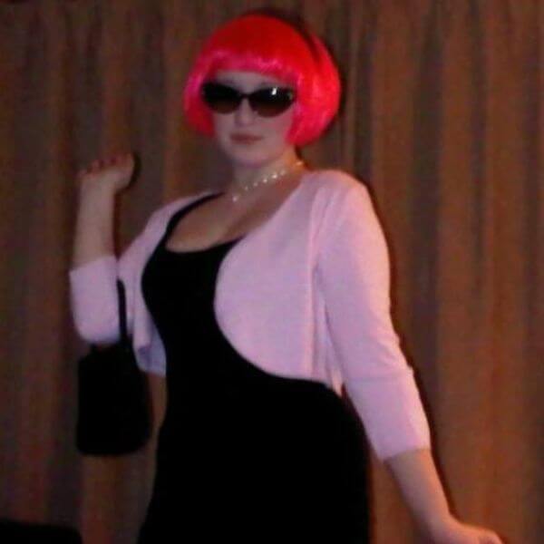 Laura, aged fourteen, dressed up as Frenchy from the movie Grease. She is posing for the camera with a handbag in the crook of her elbow. She wears a pink cardigan, bright pink wig and sunglasses.