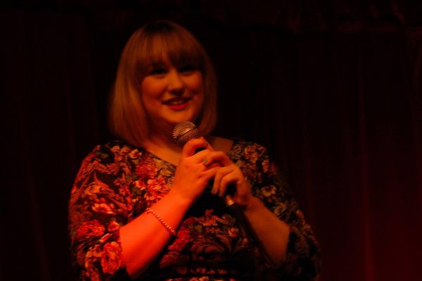 Laura, aged nineteen, performing her first ever stand-up set. Her hair is cut into a short bob with a full fringe, she is wearing a floral dress. She is smiling and holding a microphone to her mouth with both hands.