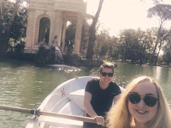 Conor and Laura on a boating lake in a park in Rome. Conor is rowing the boat and Laura is taking the picture. It is a sunny day and they are both wearing sunglasses.