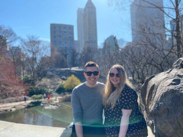 Conor and Laura sat on a bridge in Central Park, New York. The park is in the background, and behind that you can see tall New York buildings. It is a very sunny day and they are both wearing sunglasses, leaning towards one another and smiling.